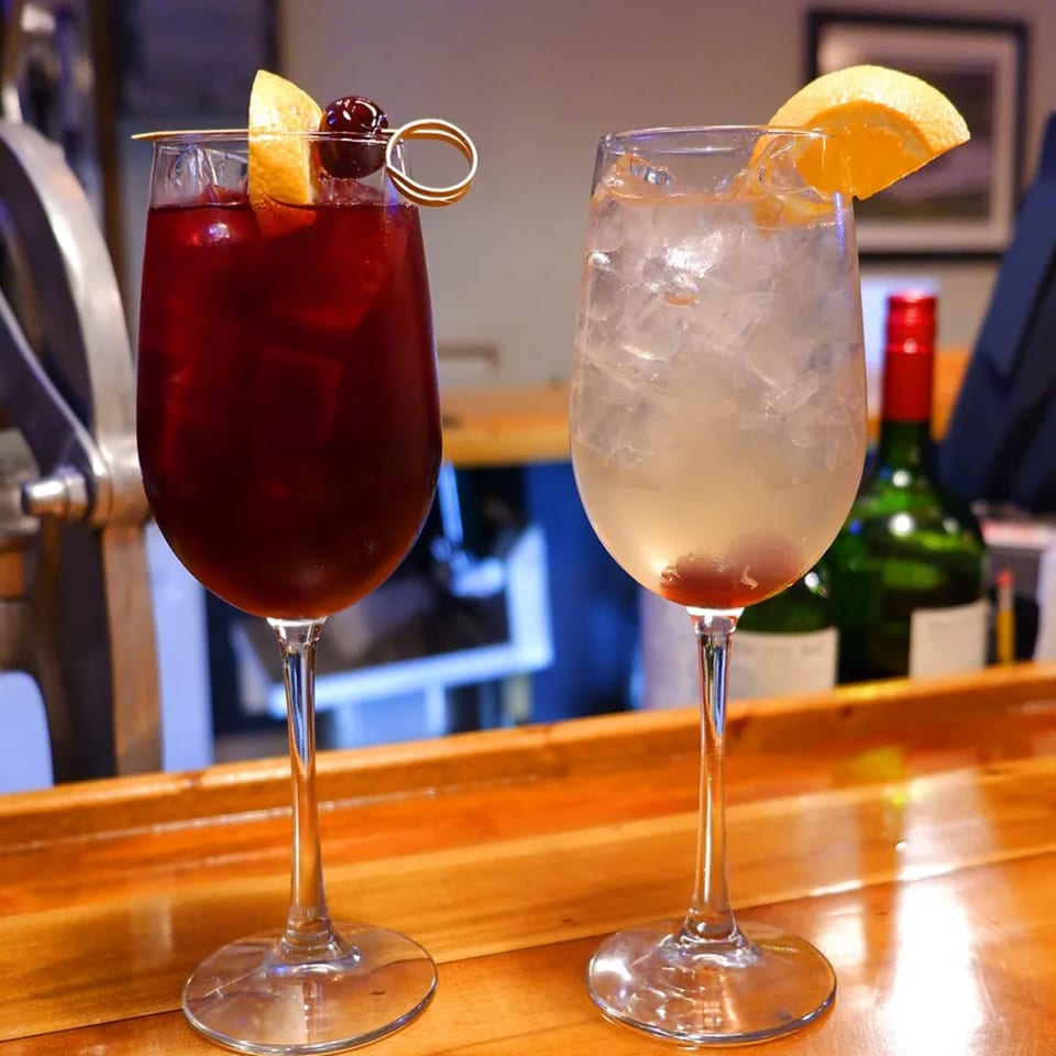 Two cocktails, one red with orange and cherry, one clear with orange, on a wooden bar.