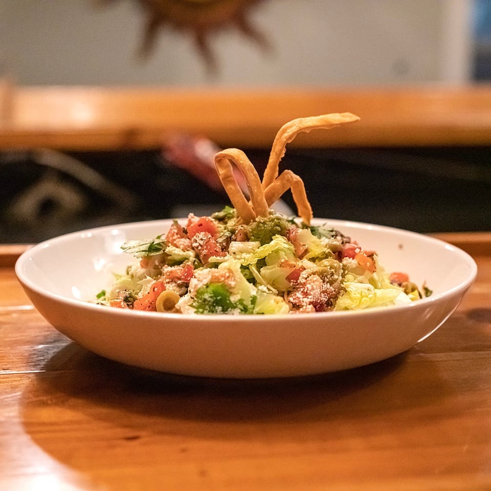 A white bowl of fresh salad with lettuce, tomatoes, olives, and crispy tortilla strips.