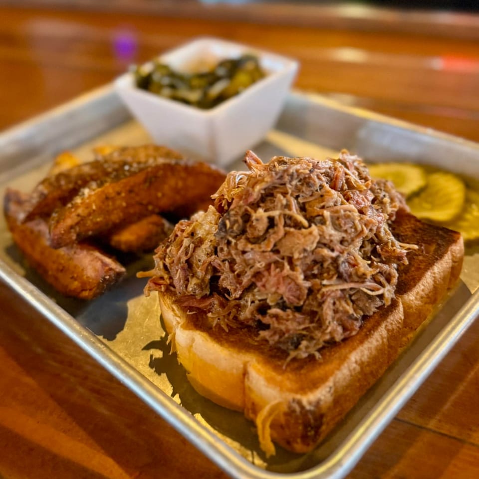 United Smokehouse pulled pork sandwich with potato wedges, collard greens, and pickles on a metal tray.