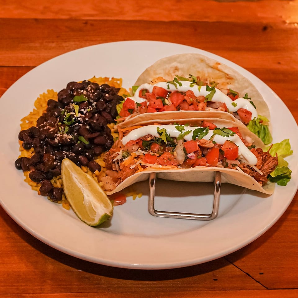 Three BBQ tacos with rice, black beans, and a lime wedge on a white plate.