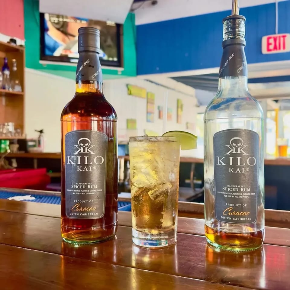 Two bottles of Kilo Kai Spiced Rum flank a rum and coke with a lime wedge on a bar.
